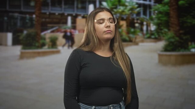 Woman with glasses closes eyes and raises hand while pointing finger in an indoor building atrium gesture of shrug and pause; contemplation calm.