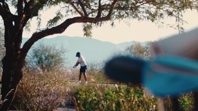 Scratch golfer swings framed through desert mesquite trees in ultra slow motion, hazy mountains beyond the branches, par 3 perfect swing on sunny summer day