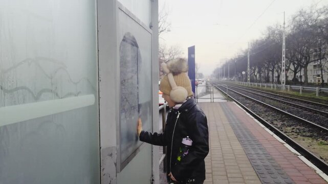 Outdoor medium shot captures a young girl in winter clothes making a funny face while wiping condensation off a foggy city map pane at an urban tram stop alongside empty railway tracks in winter.