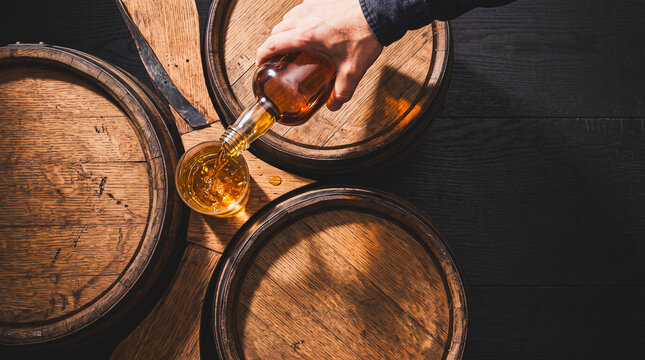 Whiskey Tasting Tradition: A hand carefully pours golden amber whiskey from a glass bottle into a crystal glass, surrounded by rustic oak barrels.