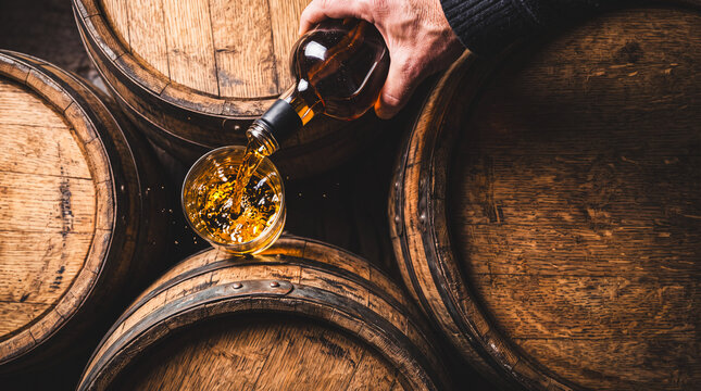 Whiskey Pouring into Glass from Bottle: A hand pours amber liquid, likely whiskey, into a short glass filled with ice, as wooden barrels surround the scene.