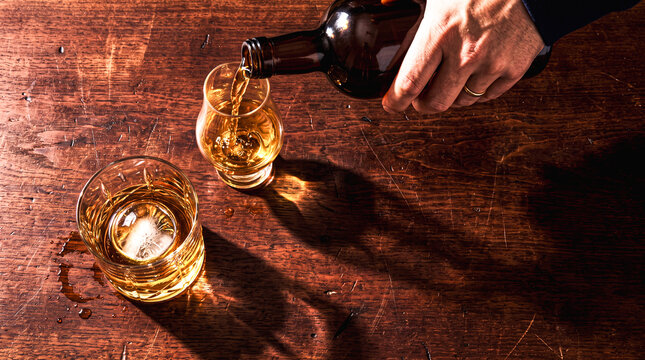 Pouring Golden Spirit: A hand pours amber liquid from a bottle into a small glass, with another glass of the same spirit on a rustic wooden table, casting long shadows.