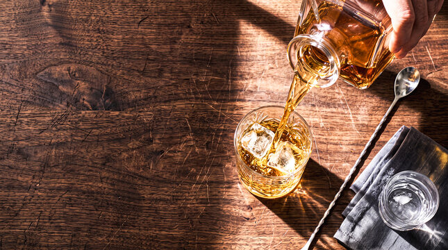 Pouring Amber Spirit: A hand pours a rich amber liquid from a decanter into a glass filled with ice cubes, set on a rustic wooden surface.