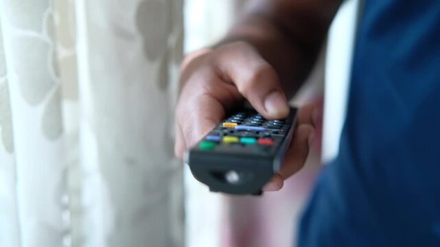 Close-up shot of a man's hand pressing buttons on a black remote control, changing channels or adjusting settings on a television screen in a bright room
