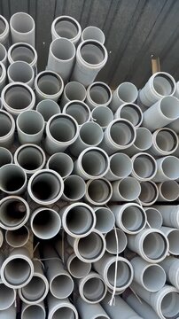 Warehouse storage scene with horizontal rows of light gray PVC pipes on wooden shelving, open ends facing camera, while upper stacks show slight outward tilt and gentle realignment