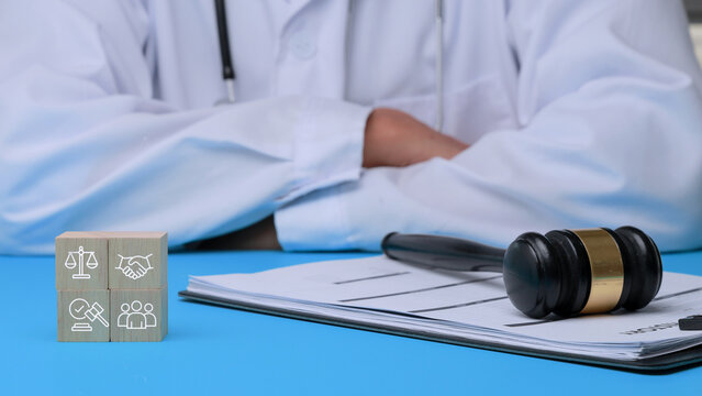Serious doctor with crossed arms faces medical malpractice law suit showing wooden block and judge gavel on blue desk inside office