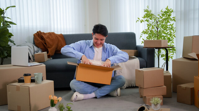 Happy young Asian man smiling while unpacking cardboard box sitting on living room floor during house move