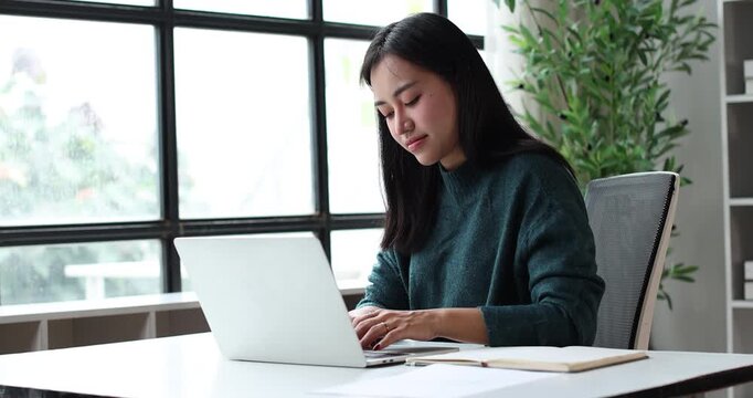 Asian woman feeling excitement and celebrating achievement while reading good news on a laptop computer, working remotely from her modern home office space