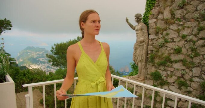 Woman holding a map on terrace with statue of Emperor Augustus overlooking Capri island and distant Faraglioni sea stacks