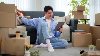 Young Asian man sitting on floor organizing new home using tablet surrounded by moving boxes © Pichsakul