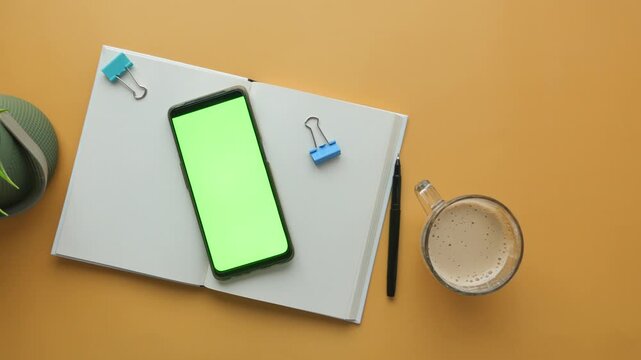 Overhead view of open notebook with green screen smartphone and coffee on yellow desk footage