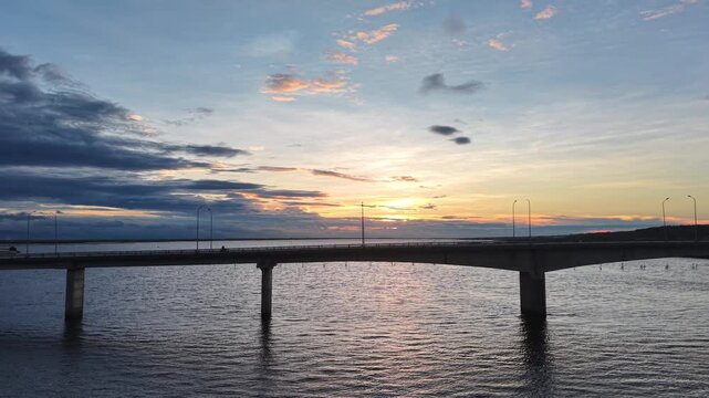 Aerial sunset view captures a bridge spanning the Hue lagoon under a dramatic sky with clouds catching the warm golden light above the calm water.