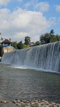 The scenic Tukad Unda Waterfall in Klungkung, Bali, Indonesia, featuring a wide cascading river dam, tropical surroundings, and a clear blue sky, creating a peaceful landscape.