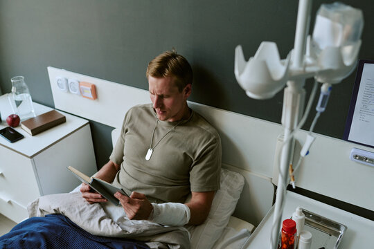 Young adult Caucasian man resting in military hospital bed, reading book with injured arm bandaged, conveying recovery and resilience. Useful for veteran healthcare campaigns