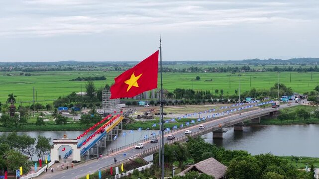 Close drone perspective of the Vietnamese national flag billowing in the wind above the Hien Luong Bridge, with green rice paddies stretching into the distance.