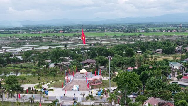 Aerial drone captures the Vietnamese national flag waving atop the tall flagpole at the Hien Luong landmark, with lush green rice paddies and rural settlement below.