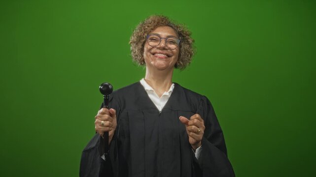 Middle-aged woman judge with curly hair holding a gavel and gesturing with hands in studio green backdrop; confidence justice.
