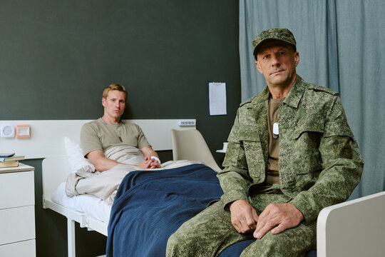 Portrait of middle aged Caucasian soldier sitting beside young adult man resting in military hospital bed, representing recovery and support. Ideal for veteran healthcare campaigns