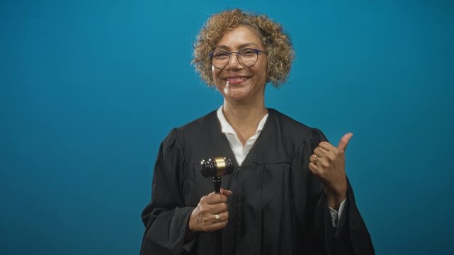 Woman judge in black robe holding gavel and giving a thumbs up gesture in studio, curly hair and glasses, smiling; confidence justice.