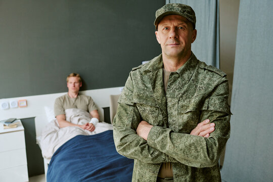 Portrait of middle aged Caucasian military man standing with arms crossed in hospital room, young adult patient recovering in background. Ideal for veteran care, military healthcare campaigns