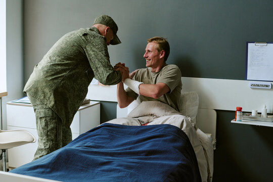 Young adult Caucasian soldier recovering in military hospital, clasping hands with fellow serviceman, showing support and rehabilitation. Ideal for veteran care, recovery campaigns