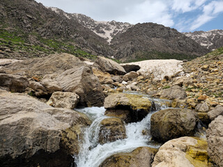 A rocky mountain stream cascades over boulders and mossy rocks near a snow cave opening, with patches of avalanche snow and rugged snow-capped peaks rising behind green hillsides. 📍 Hawraman Takht © Hawraman83