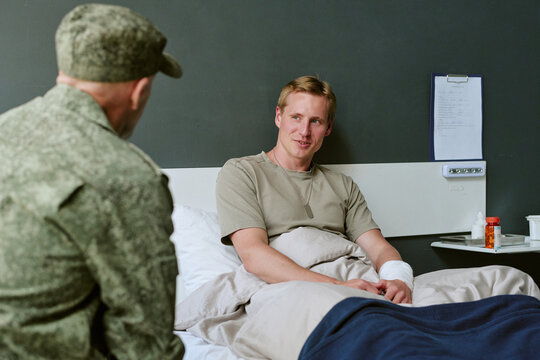 Young adult Caucasian soldier resting in military hospital bed, talking with fellow serviceman during recovery. Useful for veteran care, military medicine, rehabilitation campaigns