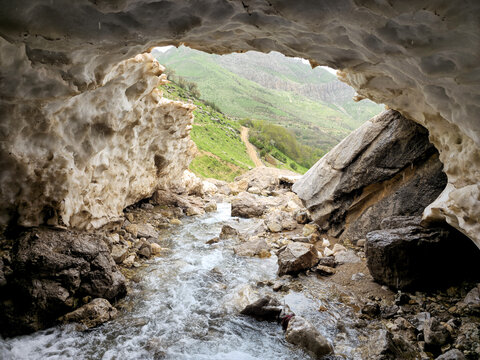 A rocky mountain stream flows out from beneath a snow cave, framed by large boulders, with a dirt trail winding along green hillsides toward mountain peaks in the background.
📍 Hawraman Takht