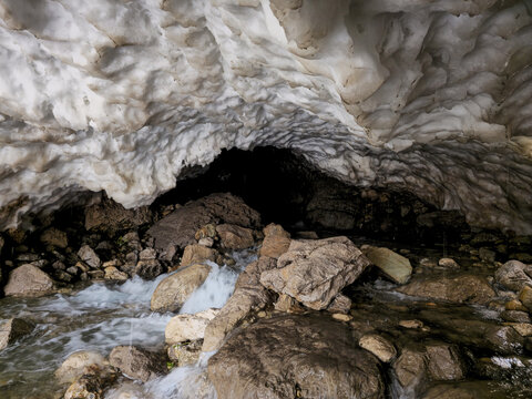 A rocky stream flows through a dark snow cave, with large boulders scattered along the water and layered snow forming an arched ceiling overhead.
📍 Hawraman Takht, Kurdistan, Iran 