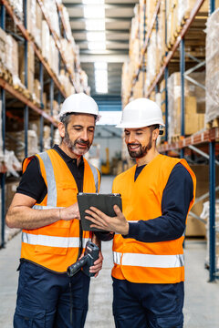 Two logistics center workers wearing safety helmets and vests using a digital tablet for inventory management and order fulfillment