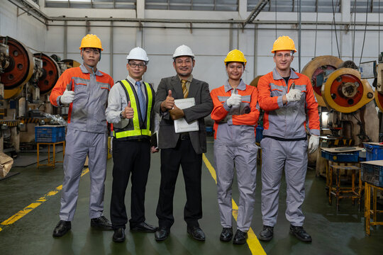 portrait multiracial manufacturing employees in safety gear with asian senior manager in suit standing in a row giving thumb up together in heavy metal factory