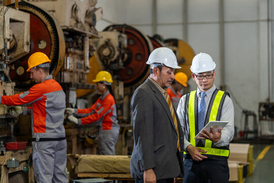 professional senior manager in suit and young male engineer wearing high-visibility vest and safety helmet using digital tablet discussing operations and quality control on a busy manufacturing plant
