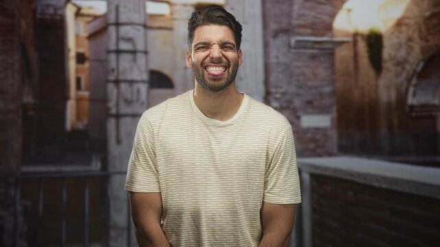 Man with beard sticks tongue out showing tongue and teeth, wearing striped beige t shirt in an old stone street; playful.