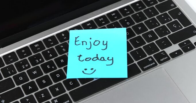 A silver laptop with a blue sticky note displaying "Enjoy today" creates a simple motivational reminder. Horizontal layout expressing optimism, productivity and mindful work habits.