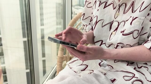 Person using a smartphone while seated by a city window. Hands hold and tap the device in a bright indoor setting. Mobile browsing and lifestyle content concept..