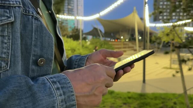 Man strolls through a city park lit with string lights in the evening, holding his phone close-up. He looks at the screen while walking beneath a warm glow. Urban leisure unfolds as he