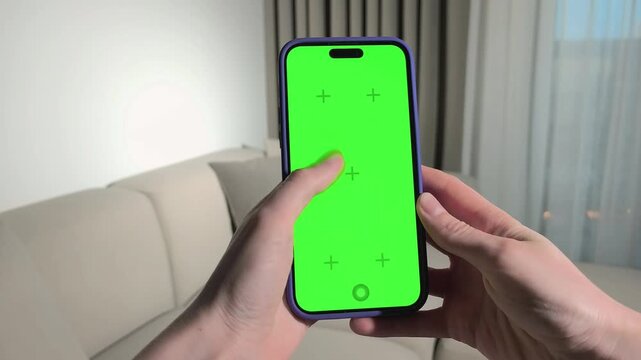 Hands holding smartphone with green screen in living room. Fingers tap and adjust the mobile device for mockup editing. App design, video overlay, and advertising template use..