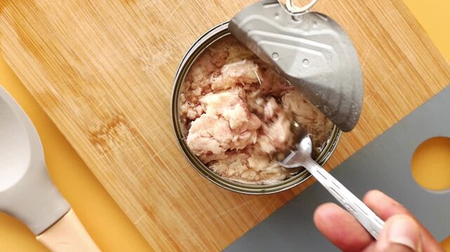 Top view of fork lifting canned tuna fish from open tin over wooden board