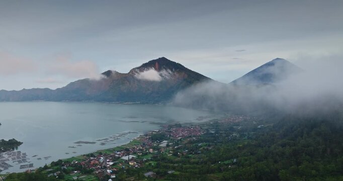 Batur caldera lake and villages resting peacefully at the base of Mount Batur and Mount Agung, low hanging mist and clouds on overcast sky. Travel and explore Bali Island, Indonesia. Aerial flight