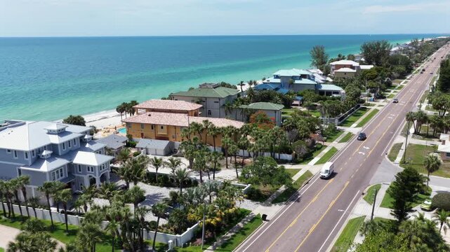 Luxury beachfront mansions line quiet coastal road in Indian Rocks Beach, Florida, turquoise Gulf waters meet white sand beach beside palm-shaded driveways plus pools. Aerial drone flyover shot