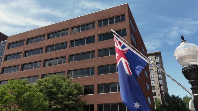 Australian flag flies below clear blue sky beside tall brick office building corner in American downtown, ornate vintage streetlamp stands near trees with apartment tower rising behind. low angle