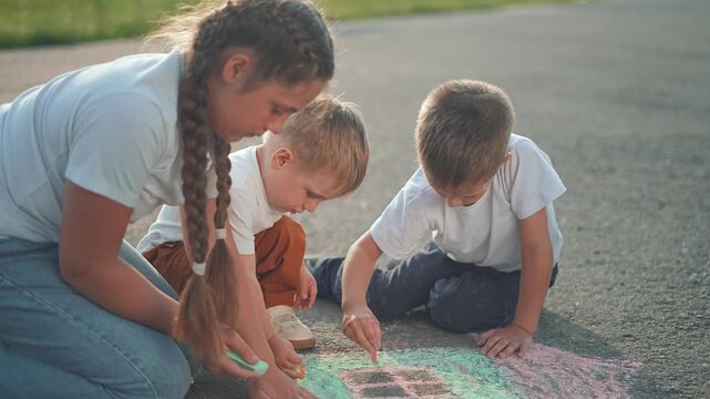Boy grabs chalk from ground. Children play together drawing shapes on city street. Chalk lines swirl in playful mess. Toddler joins fun. Kids create drawing patterns. Children chalk on pavement.
