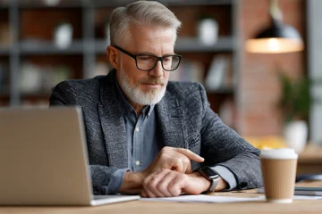 Senior businessman sitting at office desk with coffee and laptop checking his smartwatch © Delphotostock