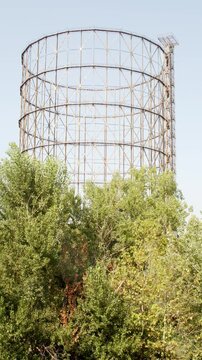 A large industrial gasometer framework rises high into the clear sky behind lush green tree tops, showcasing a blend of urban metal architecture and vibrant natural foliage on a bright, sunny day.