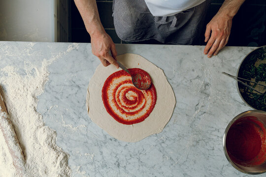 A pizzaiolo spreads red sauce onto a pizza base with a spoon.