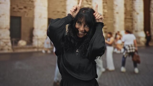 Woman makes horns with fingers and smiles, hands raised by her head on a cobblestone street before a historic stone building; playful mischief.