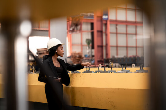 African Female Engineer in Blue Hard Hat Inspecting Industrial Equipment at Steel Factory. Female Professional Conducting Quality Control in Manufacturing.