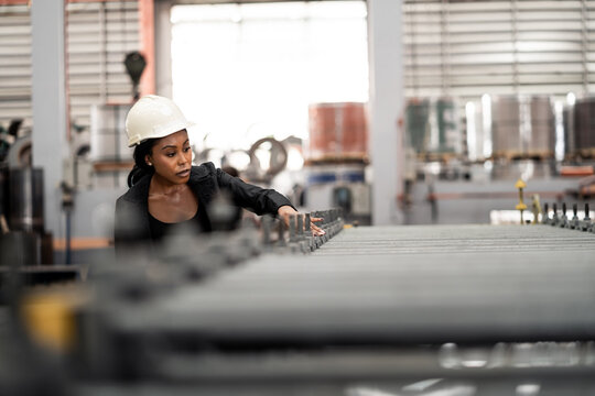 African Female Engineer in Blue Hard Hat Inspecting Industrial Equipment at Steel Factory. Female Professional Conducting Quality Control in Manufacturing.