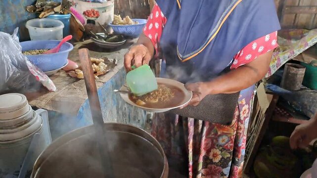 Indonesian street food vendor serving hot green bean broth with meat, traditional Madura cuisine. Steaming hot smoke rising from pot, preparing customer order at local warung.