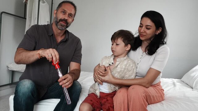 A boy, his mother, and his father sit together on the bed, playing with fun toys and holding a bubble blower at home. A family-themed concept.
